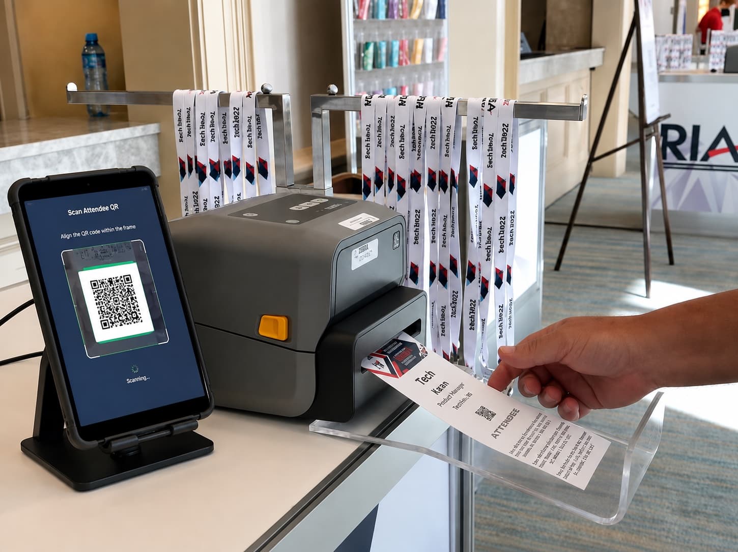A Zebra desktop printer feeding out a finished attendee badge at a check-in desk, next to a tablet scanning a guest's QR code and a rack of lanyards